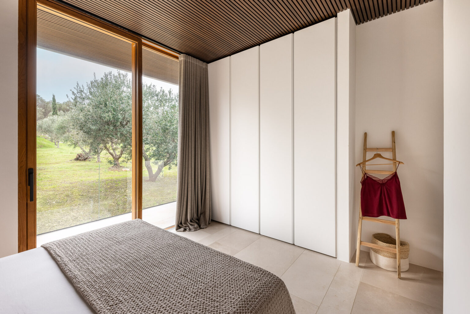 Modern bedroom featuring large glass windows with views of olive trees, minimalist white cabinetry, a cozy textured blanket on the bed, and a stylish wooden ladder displaying a red garment and woven basket. | Only on SopranoVillas