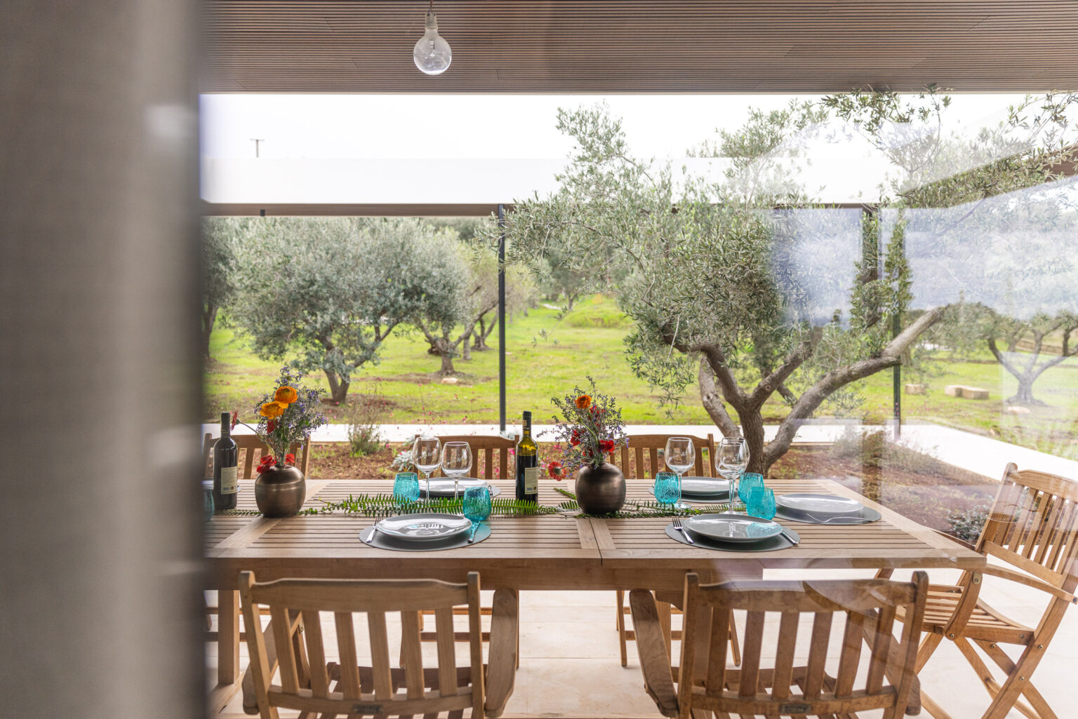 Dining area with a wooden table set for a meal, featuring blue glassware, plates, and a vase of flowers, overlooking an olive grove. Natural light enhances the inviting atmosphere of the outdoor space. | Only on SopranoVillas