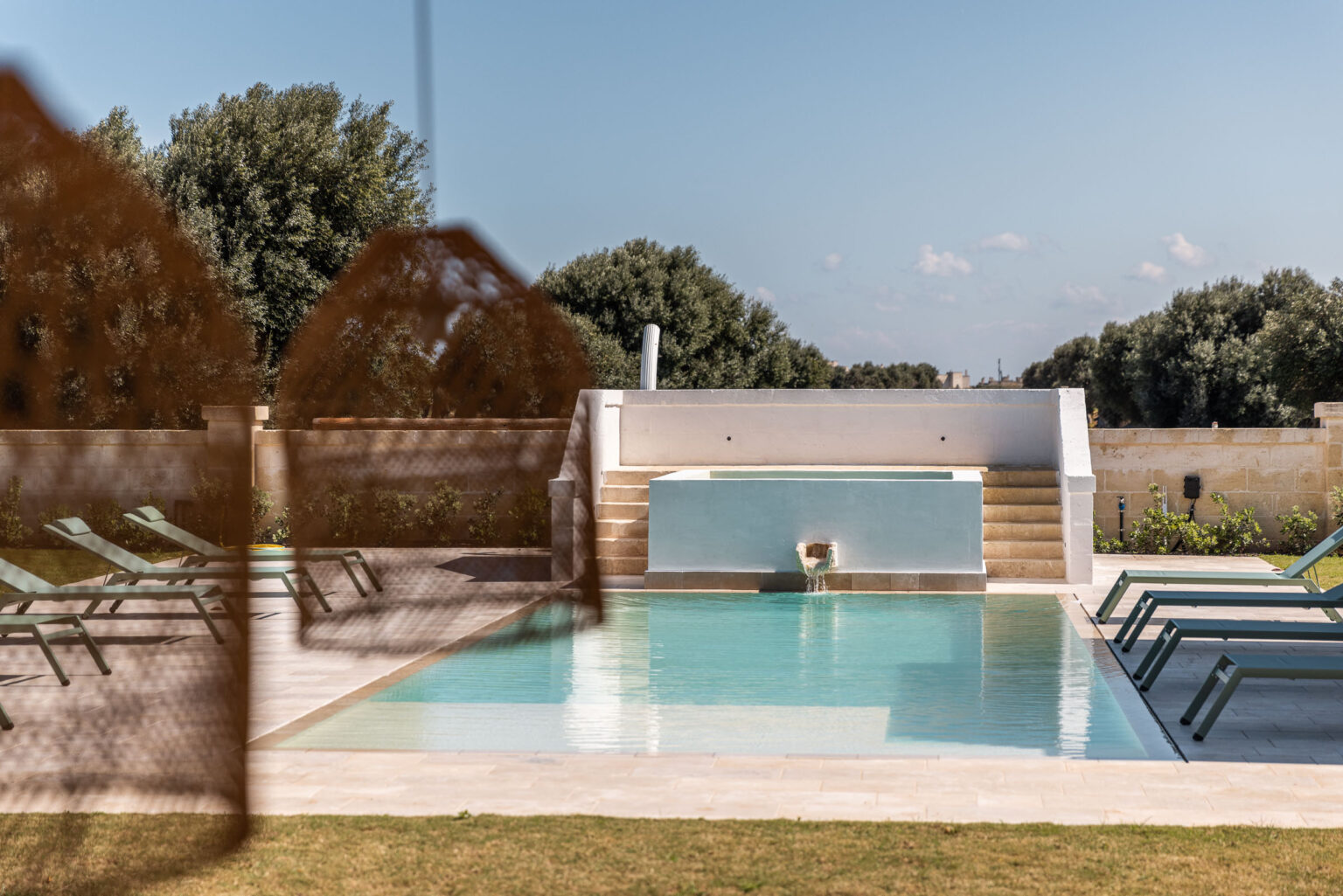 Modern swimming pool surrounded by green lounge chairs and a serene garden, featuring a stone wall and lush trees in the background under a clear blue sky. Ideal for relaxation and leisure. | Only on SopranoVillas