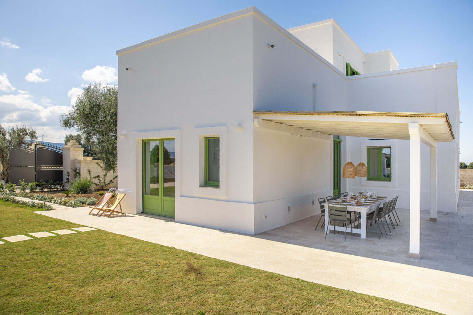 Modern white house with green accents and a spacious outdoor dining area, surrounded by a well-maintained lawn and sunny blue skies. Ideal for relaxing and entertaining. | Only on SopranoVillas