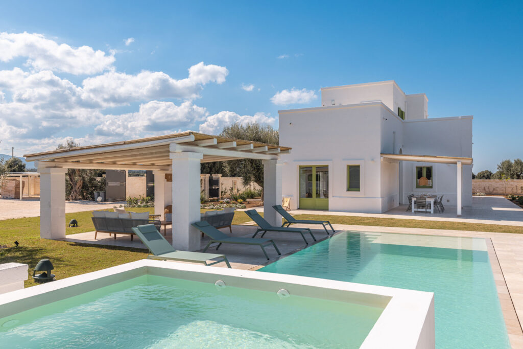 Modern villa with swimming pool and outdoor seating area, surrounded by lush greenery and blue sky. The property features a stylish design with large windows and a spacious patio, ideal for relaxation and entertaining. | Only on SopranoVillas
