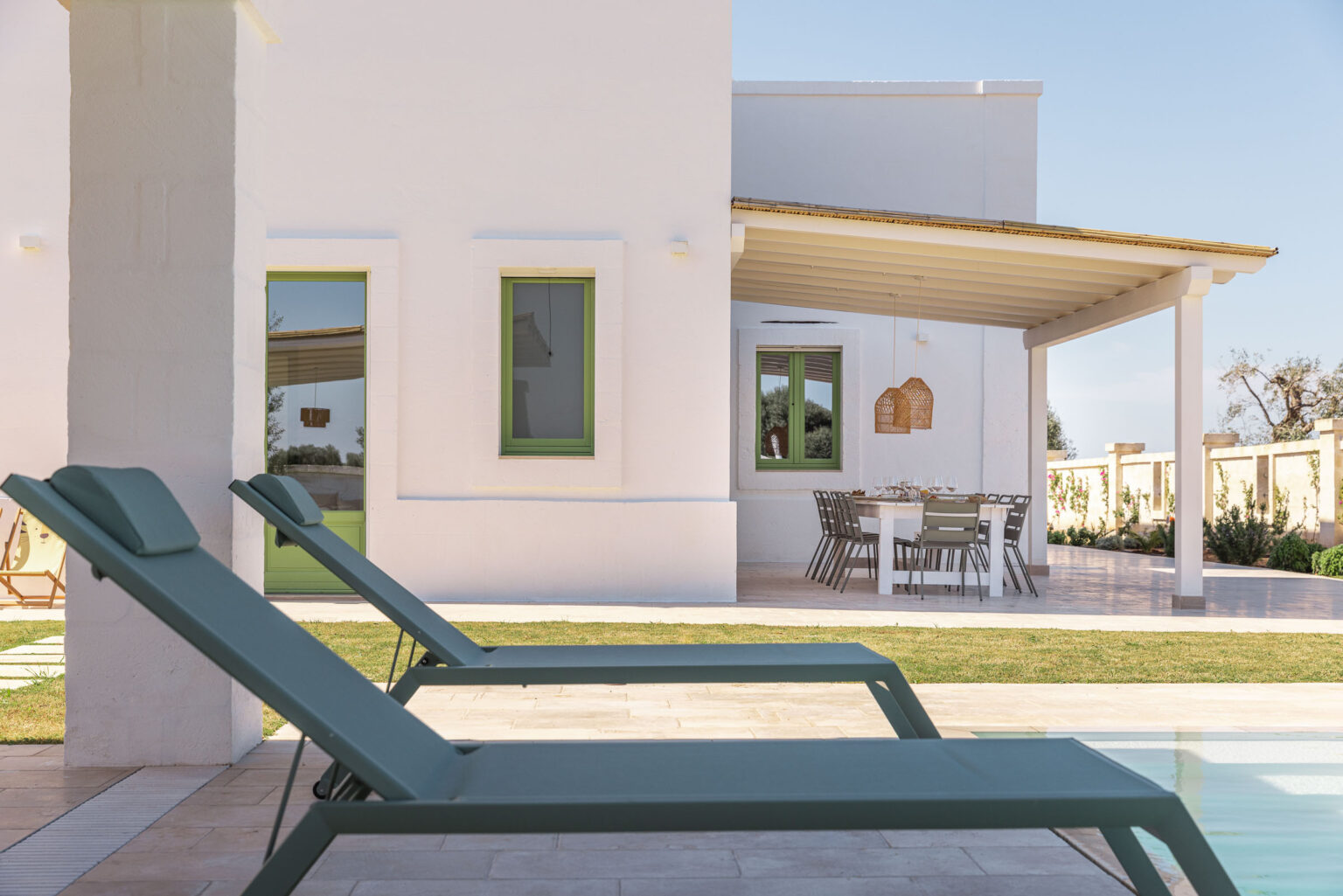 Modern outdoor lounge chairs beside a swimming pool in front of a stylish white house with green accents and a shaded patio area. Ideal for relaxation and outdoor living. | Only on SopranoVillas
