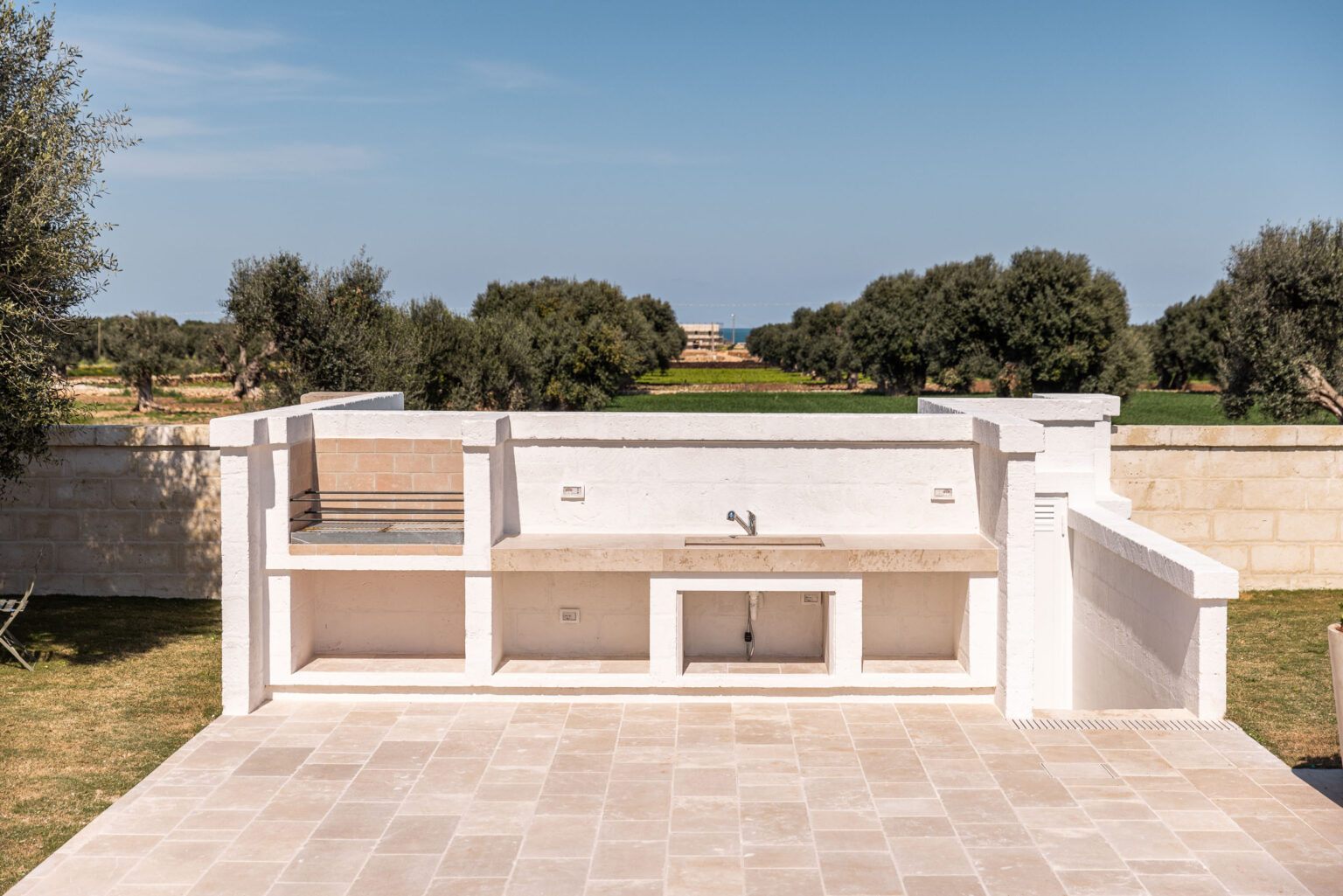 Outdoor kitchen setup featuring a stone countertop, sink, and built-in grill, surrounded by lush greenery and olive trees, ideal for alfresco dining and entertaining. | Only on SopranoVillas