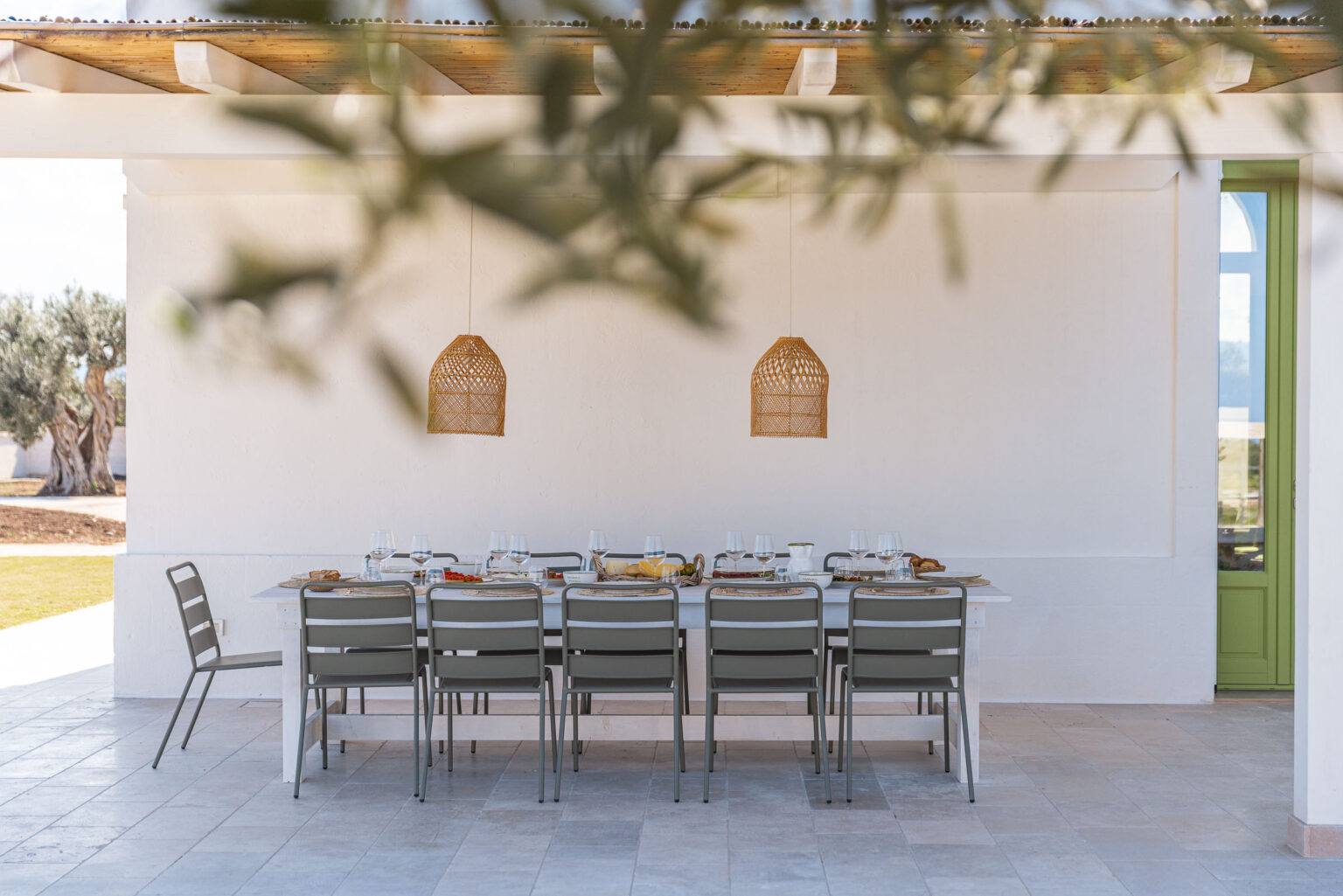 Outdoor dining area featuring a long table set with plates and glasses, surrounded by modern chairs. Two woven pendant lights hang above the table against a white wall, with an olive tree visible in the background. The setting is bright and inviting, perfect for al fresco meals. | Only on SopranoVillas