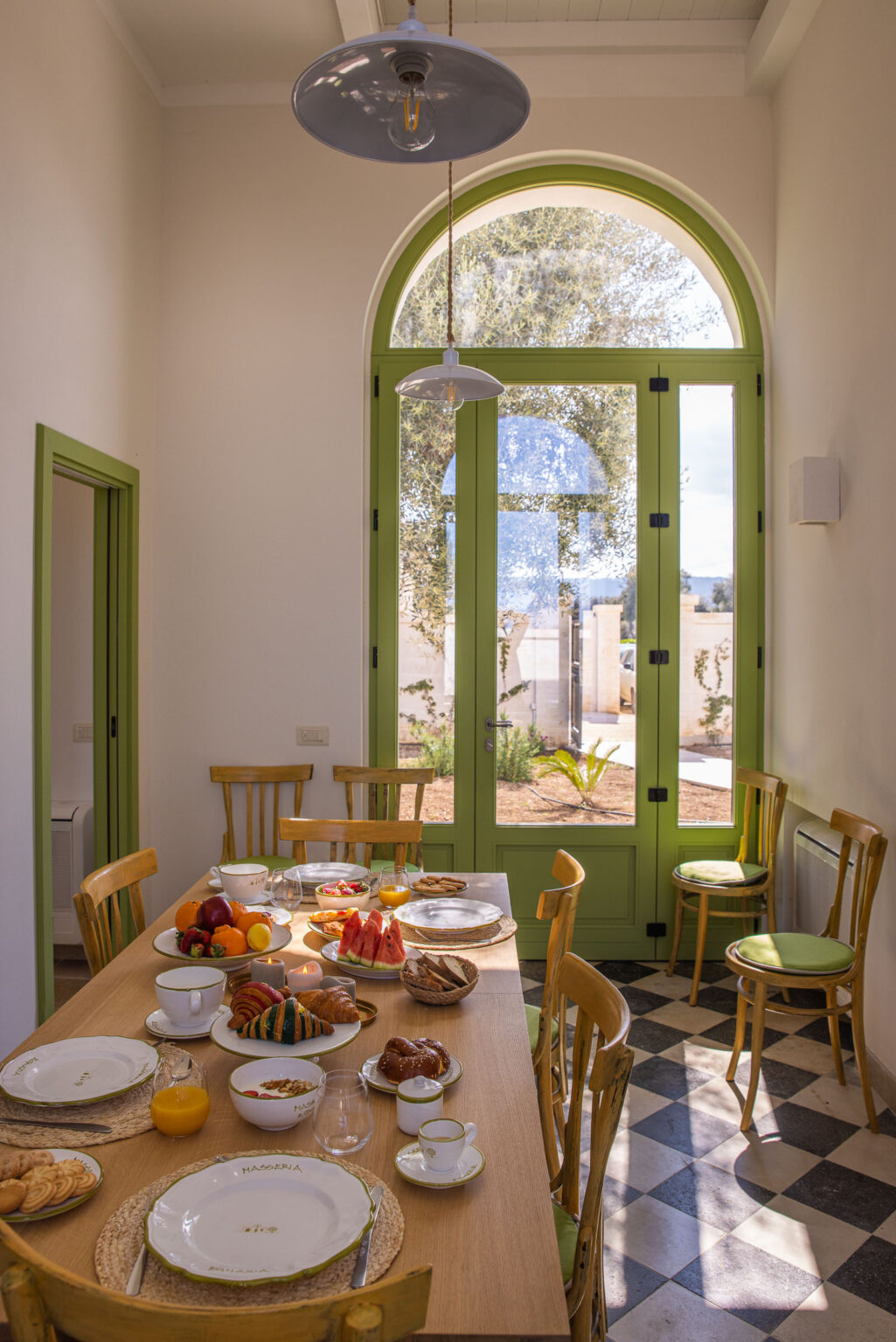 Bright dining area featuring a wooden table set with a variety of fresh fruits and plates, surrounded by green-framed windows that provide a view of the outdoor garden. | Only on SopranoVillas