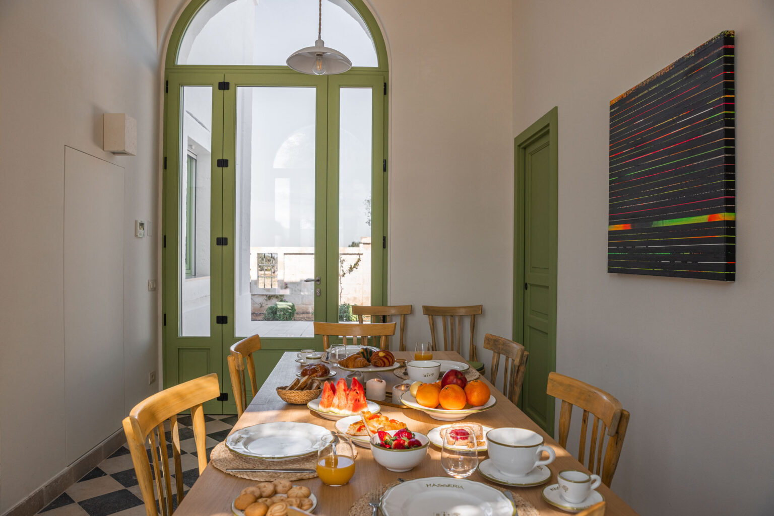Bright dining area featuring a wooden table set with a variety of breakfast foods, including fruits, pastries, and drinks. Large green-framed windows allow natural light to fill the room, complemented by a colorful abstract painting on the wall. The space exudes a warm and inviting atmosphere, perfect for enjoying a morning meal. | Only on SopranoVillas