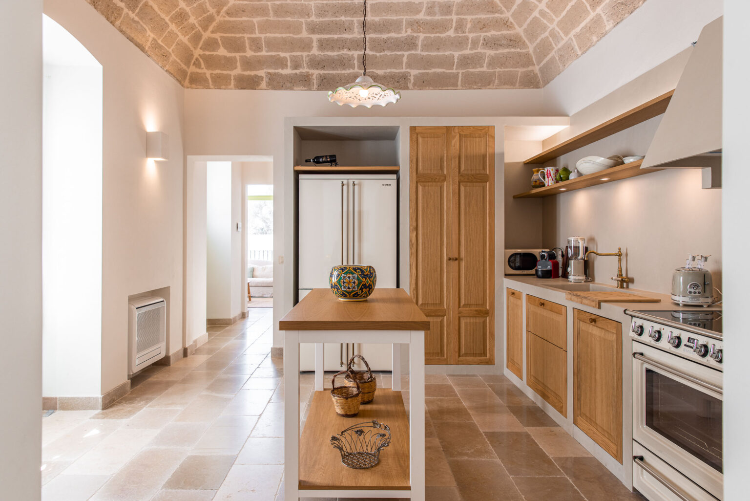 Bright and modern kitchen featuring a high arched ceiling, wooden cabinetry, and a central island with decorative bowls. The kitchen is equipped with stainless steel appliances, including a refrigerator and oven, complemented by warm lighting and natural stone flooring. | Only on SopranoVillas