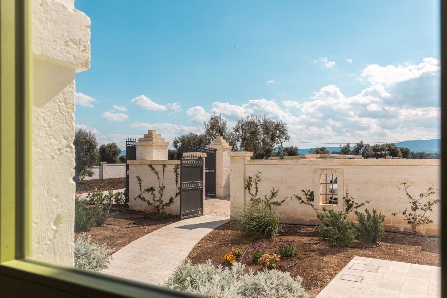 Scenic view from a window showcasing a landscaped garden with a pathway leading to a rustic stone wall and an open gate, under a bright blue sky with fluffy clouds. | Only on SopranoVillas
