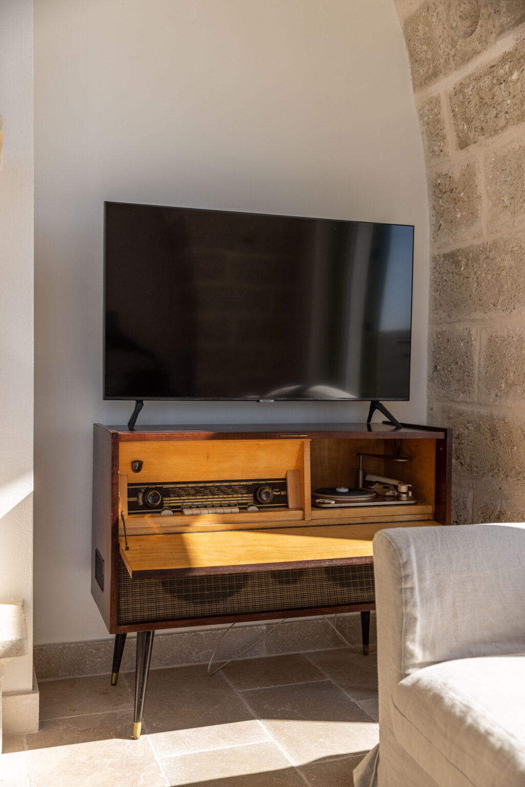 Modern living room setup featuring a flat-screen TV mounted above a vintage wooden media console with a record player and radio. The console has a sleek design with a warm wood finish, complemented by a light-colored sofa and natural stone wall in the background. | Only on SopranoVillas