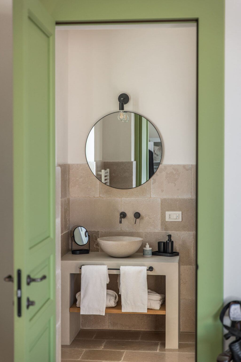 Modern bathroom interior featuring a round mirror, stylish sink, and green door. The space includes stone walls, black fixtures, and neatly arranged towels, creating a minimalist and elegant atmosphere. | Only on SopranoVillas