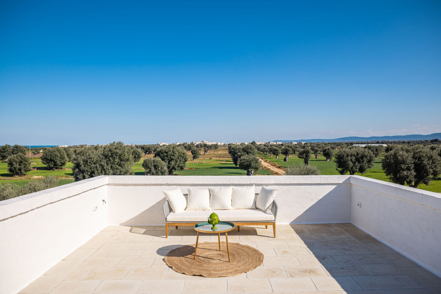 Modern rooftop terrace with a cozy sofa and coffee table, overlooking lush green fields and olive trees under a clear blue sky. Perfect for outdoor relaxation and enjoying scenic views. | Only on SopranoVillas
