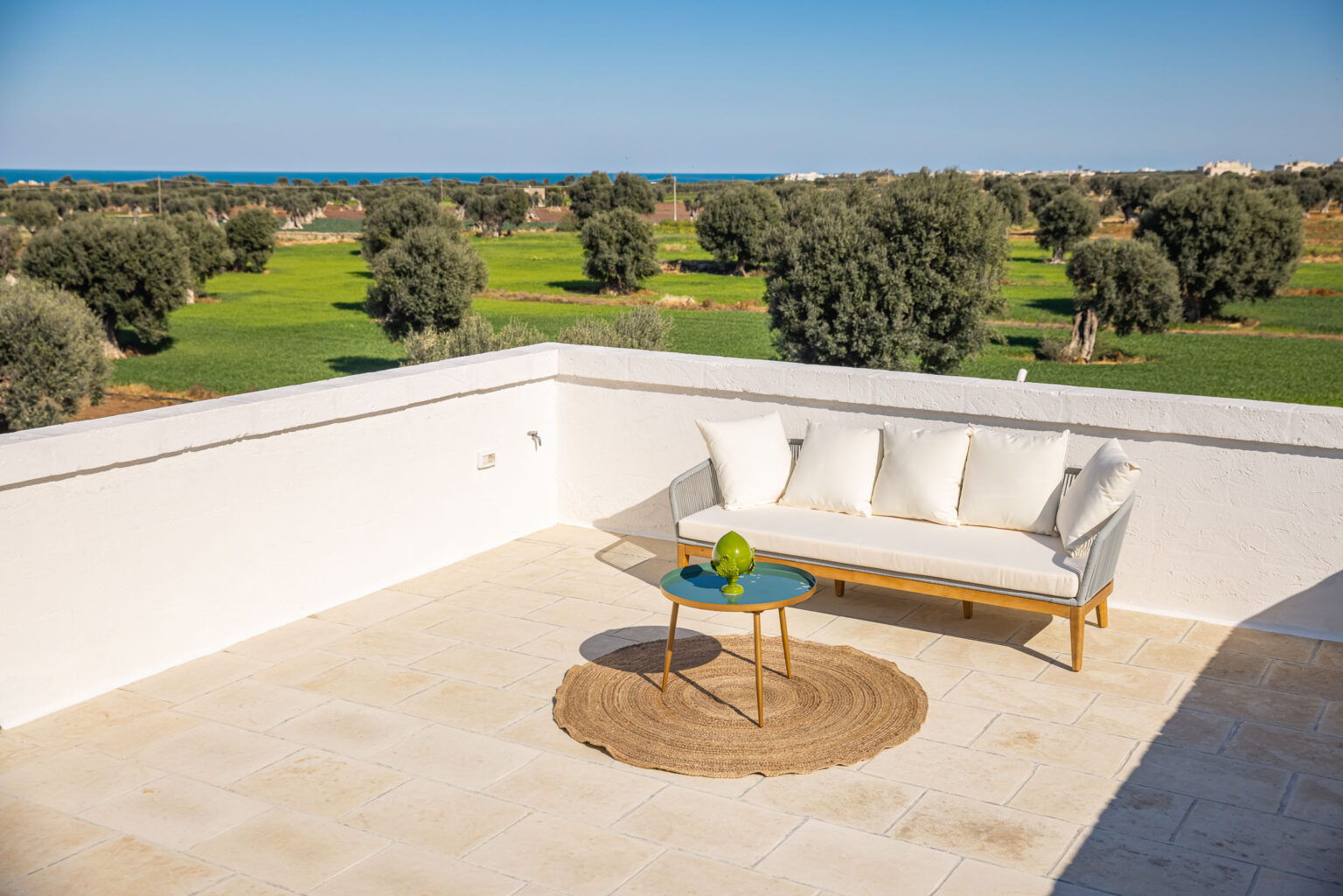 Stylish outdoor terrace featuring a comfortable white sofa with plush cushions, a round jute rug, and a modern green accent table, overlooking lush green fields and the ocean in the background. Perfect for relaxation and enjoying scenic views. | Only on SopranoVillas