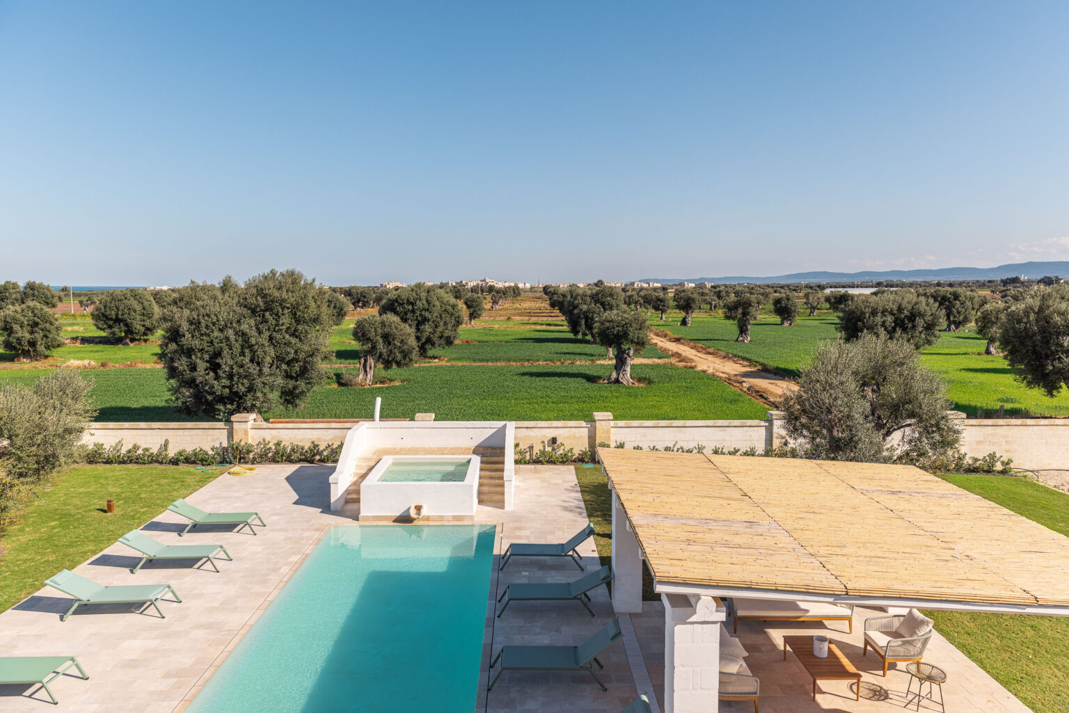 Aerial view of a modern villa with a swimming pool surrounded by olive trees and lush green fields under a clear blue sky. | Only on SopranoVillas