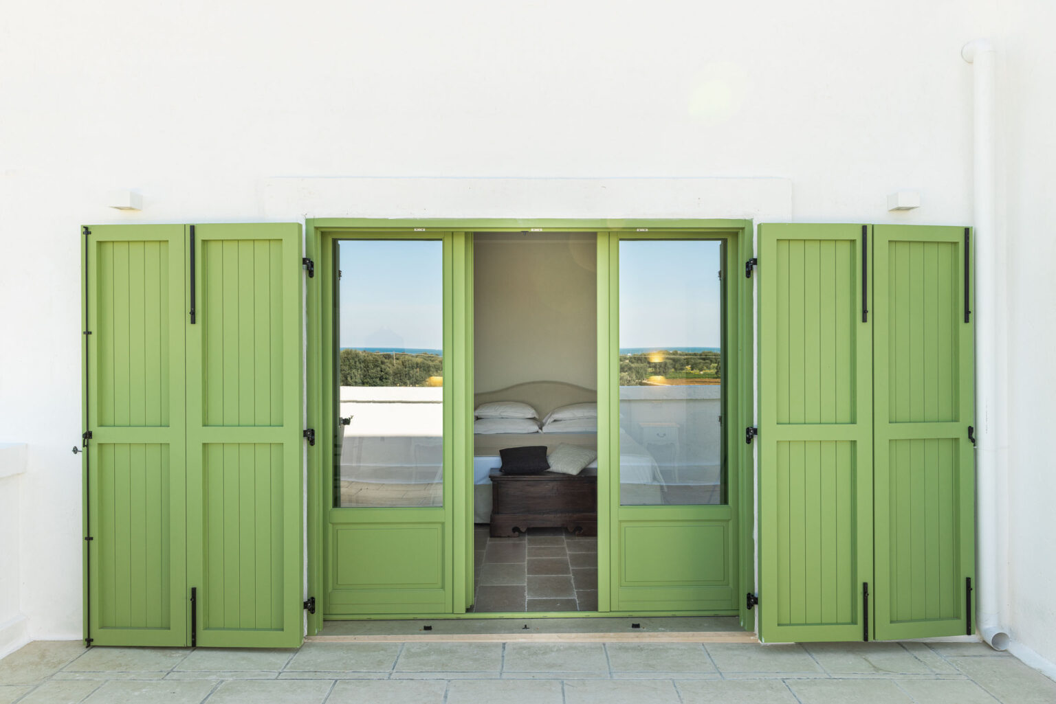 Green wooden shutters open to reveal a bright bedroom with a bed and decorative pillows, framed by a view of the outdoors. The scene captures a serene and inviting atmosphere, ideal for relaxation. | Only on SopranoVillas