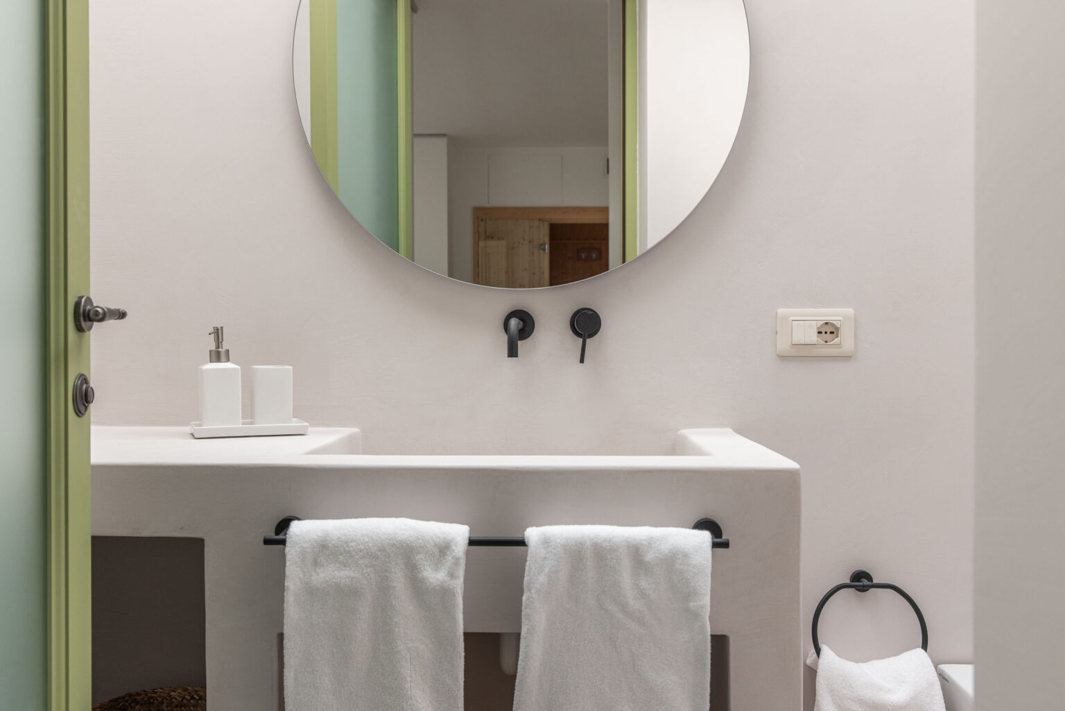 Modern bathroom featuring a minimalist design with a round mirror, black faucets, and a white sink. Soft white towels hang on a black towel rack, complemented by a stylish green door and neutral wall tones. Ideal for showcasing contemporary interior decor. | Only on SopranoVillas