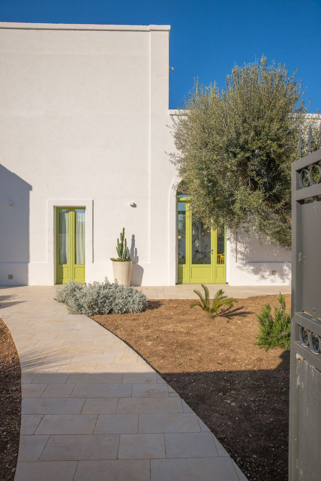 Modern white house exterior with vibrant green doors, surrounded by a landscaped garden featuring cacti, olive trees, and decorative shrubs. A curved stone pathway leads to the entrance, under a clear blue sky. | Only on SopranoVillas