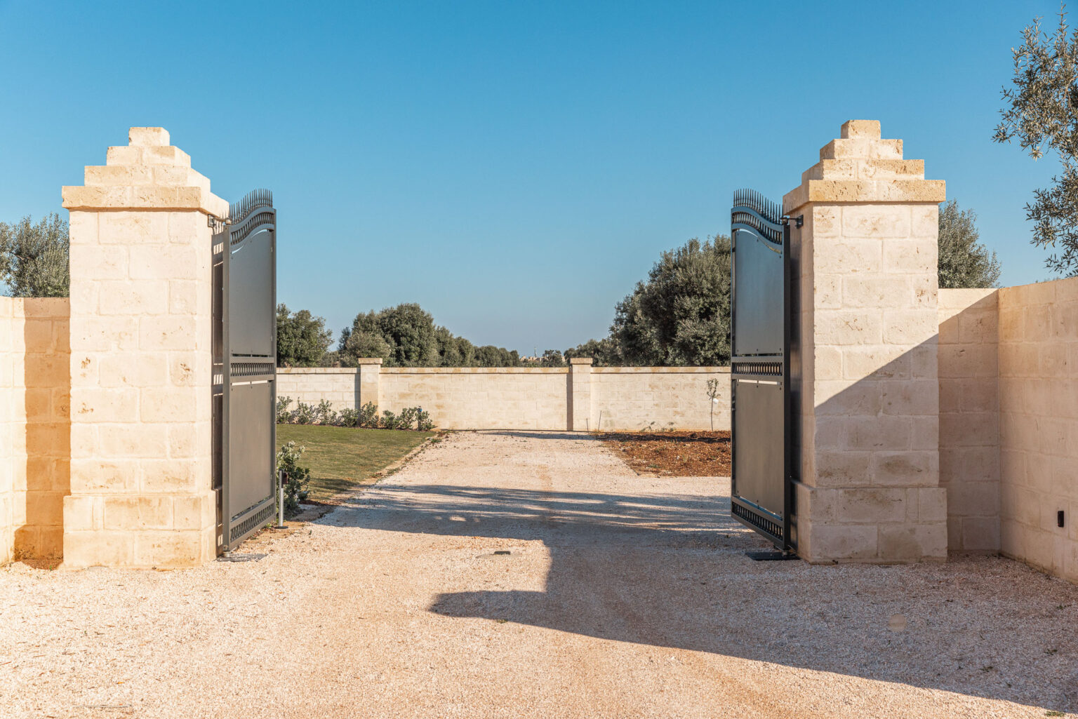 Open stone gates leading to a gravel driveway, framed by olive trees under a clear blue sky, showcasing a serene entrance to a property. | Only on SopranoVillas