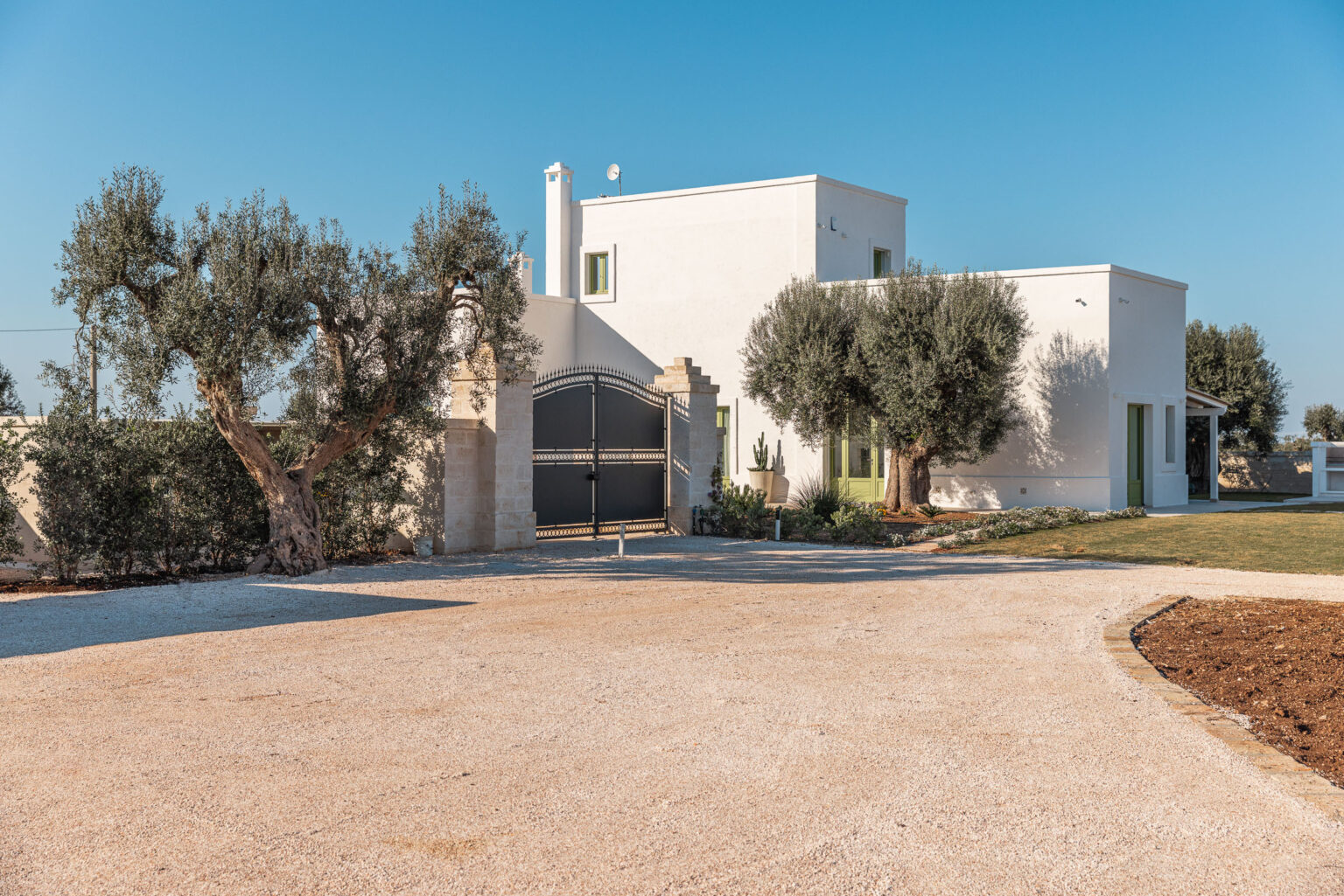 Modern white villa with olive trees, gravel driveway, and clear blue sky, showcasing contemporary architecture in a serene landscape. | Only on SopranoVillas