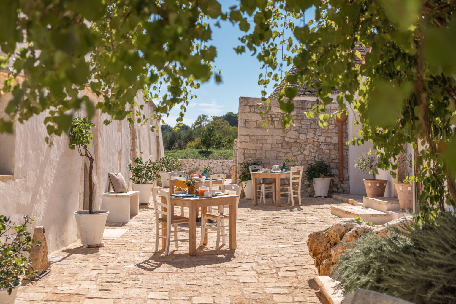 Charming outdoor dining area with rustic wooden tables set for a meal, surrounded by lush greenery and white stone walls, perfect for a serene dining experience in a Mediterranean setting. | Only on SopranoVillas
