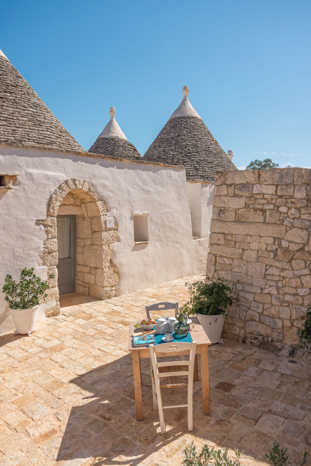 Tranquil outdoor dining area featuring a wooden table set with dishes, surrounded by traditional whitewashed trulli houses under a clear blue sky. | Only on SopranoVillas