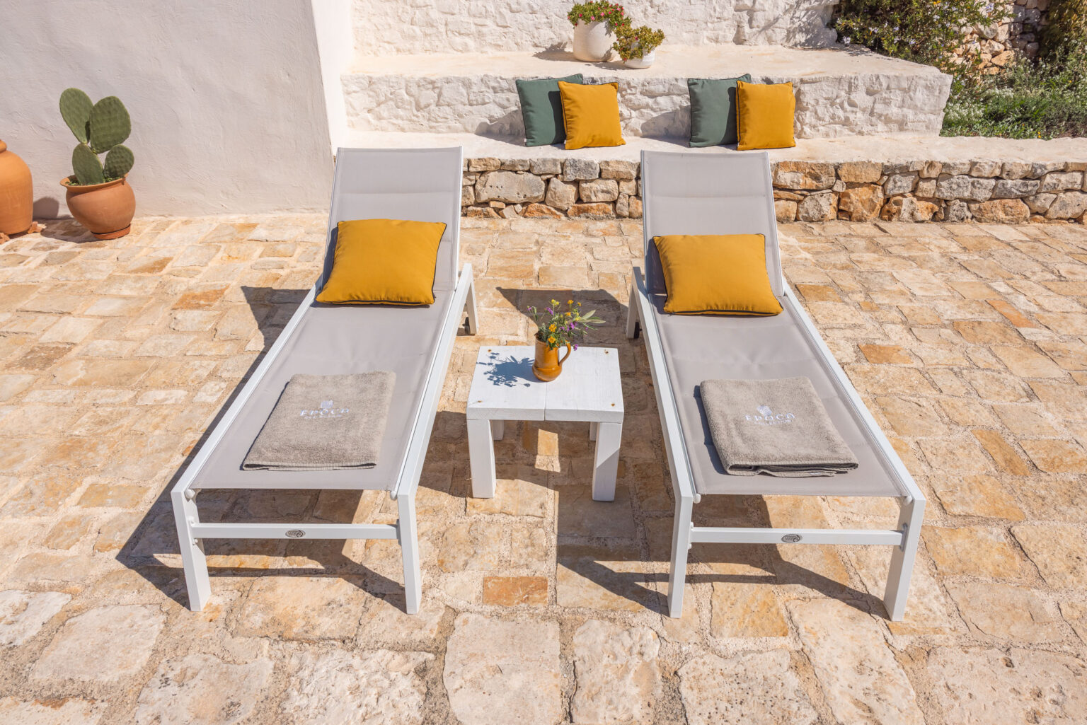 Two modern grey lounge chairs with yellow and green cushions sit on a sunlit stone patio, accompanied by a small white table holding a vase of fresh flowers. A potted cactus and decorative planters are visible in the background, creating a serene outdoor relaxation space. | Only on SopranoVillas