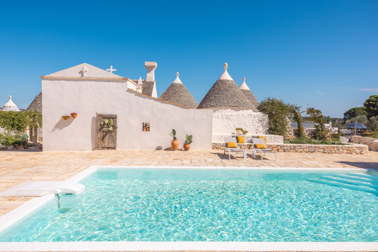 Tranquil vacation villa featuring traditional trullo architecture, a sparkling swimming pool, and a sunny outdoor seating area surrounded by lush greenery. Ideal for relaxation in a picturesque setting. | Only on SopranoVillas