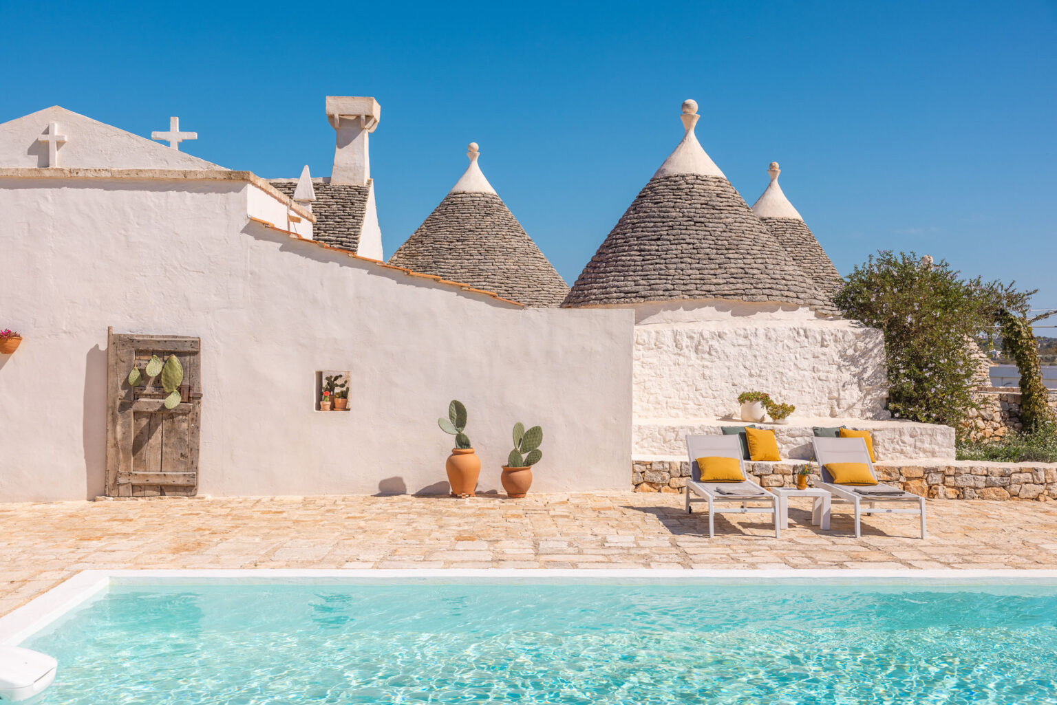 Tranquil outdoor scene featuring a swimming pool, traditional trulli houses with conical roofs, and a sunbathing area with lounge chairs and potted plants. The clear blue sky enhances the serene atmosphere of this picturesque Italian villa. | Only on SopranoVillas
