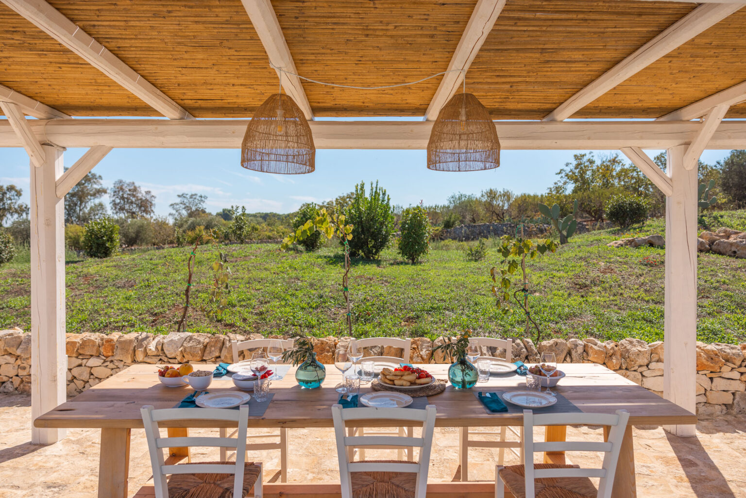 Outdoor dining area featuring a wooden table set for a meal, surrounded by lush greenery and stone walls. The scene includes decorative blue glass vases, plates, and fresh fruit, under a bamboo roof with woven pendant lights, creating a serene and inviting atmosphere for al fresco dining. | Only on SopranoVillas
