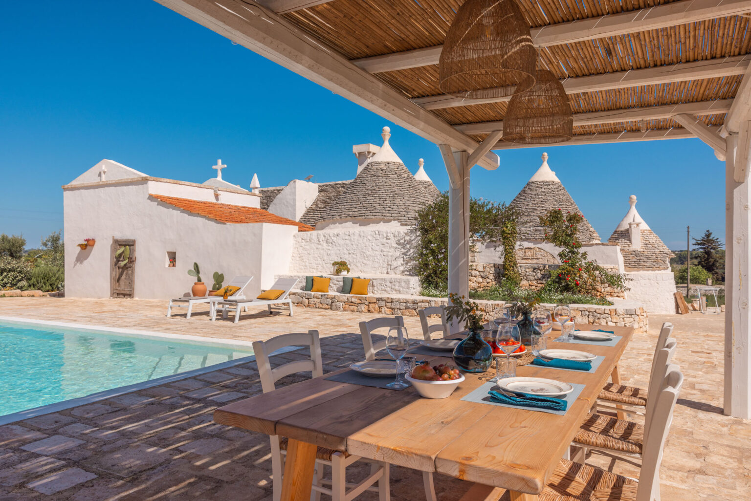 Tranquil outdoor dining area with a wooden table set for a meal, overlooking a refreshing swimming pool and traditional trulli houses in a sunny Italian landscape. | Only on SopranoVillas