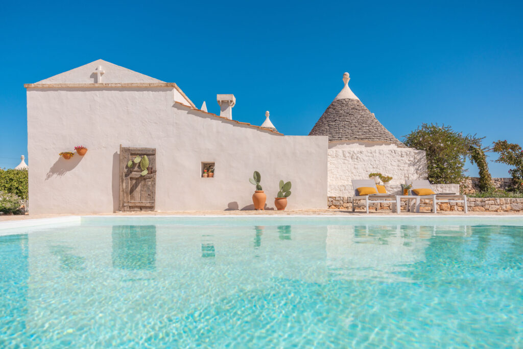 Tranquil poolside view of a traditional whitewashed trullo house in Puglia, Italy, featuring a wooden door, terracotta planters with cacti, and a clear blue sky. Ideal for vacation rentals and summer getaways. | Only on SopranoVillas