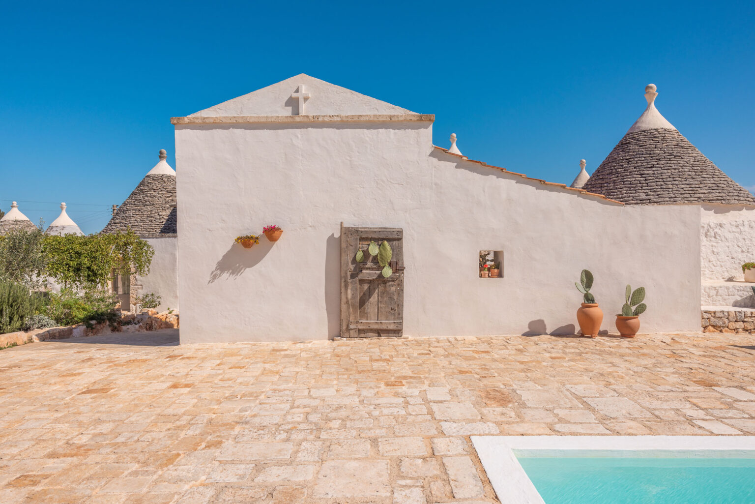 Charming trullo house in Puglia, Italy, featuring a rustic wooden door, potted flowers, and cacti, set against a clear blue sky and traditional stone flooring. | Only on SopranoVillas