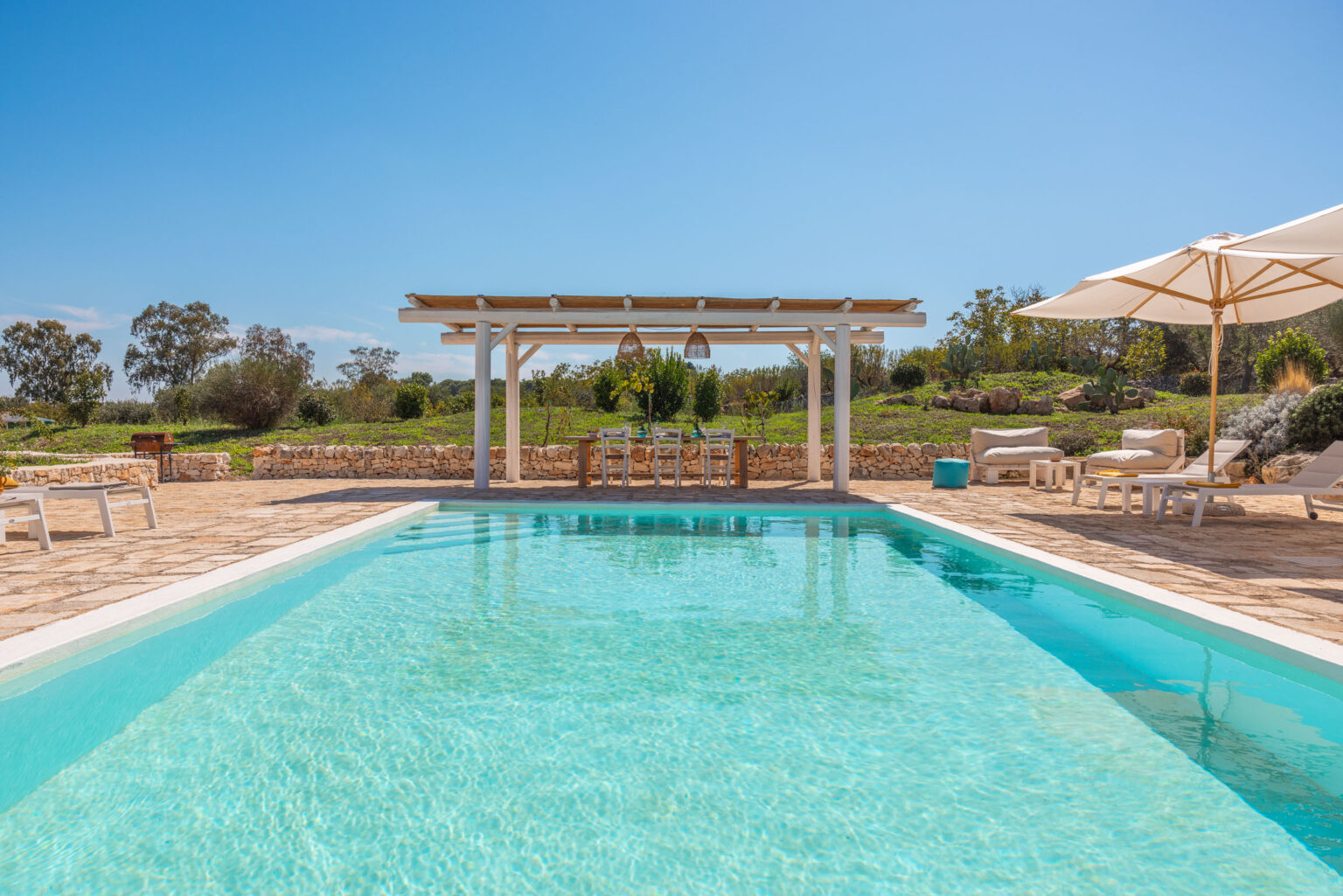 Luxurious outdoor swimming pool with crystal-clear water, surrounded by sun loungers and a shaded dining area under a wooden pergola. Lush green landscape in the background, ideal for relaxation and outdoor gatherings. | Only on SopranoVillas