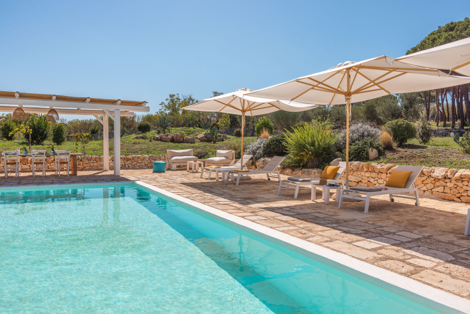 Luxurious outdoor pool area featuring comfortable sun loungers and shaded seating under large umbrellas, surrounded by lush greenery and stone walls, ideal for relaxation and leisure. | Only on SopranoVillas