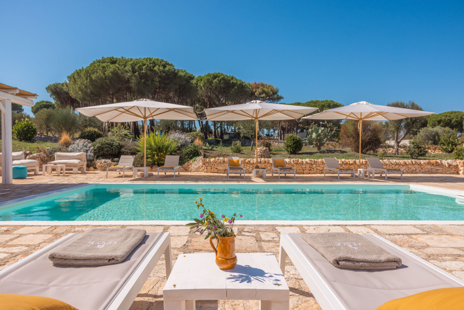 Luxurious outdoor pool area with sun loungers and large umbrellas, surrounded by lush greenery and stone walls, perfect for relaxation and leisure in a sunny setting. | Only on SopranoVillas