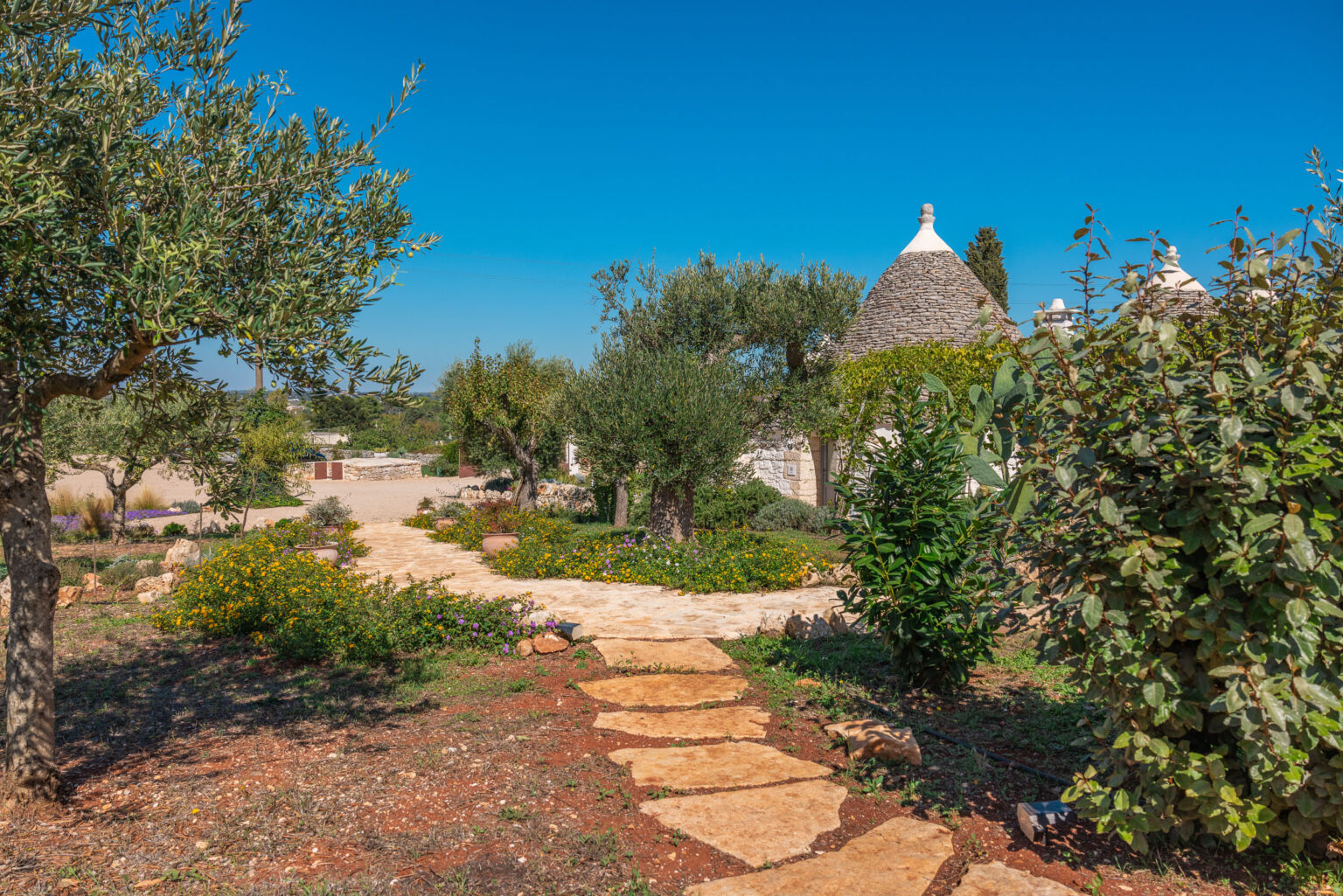 A scenic pathway lined with vibrant flowers and greenery leads to traditional trulli houses under a clear blue sky in a tranquil Italian countryside setting. | Only on SopranoVillas