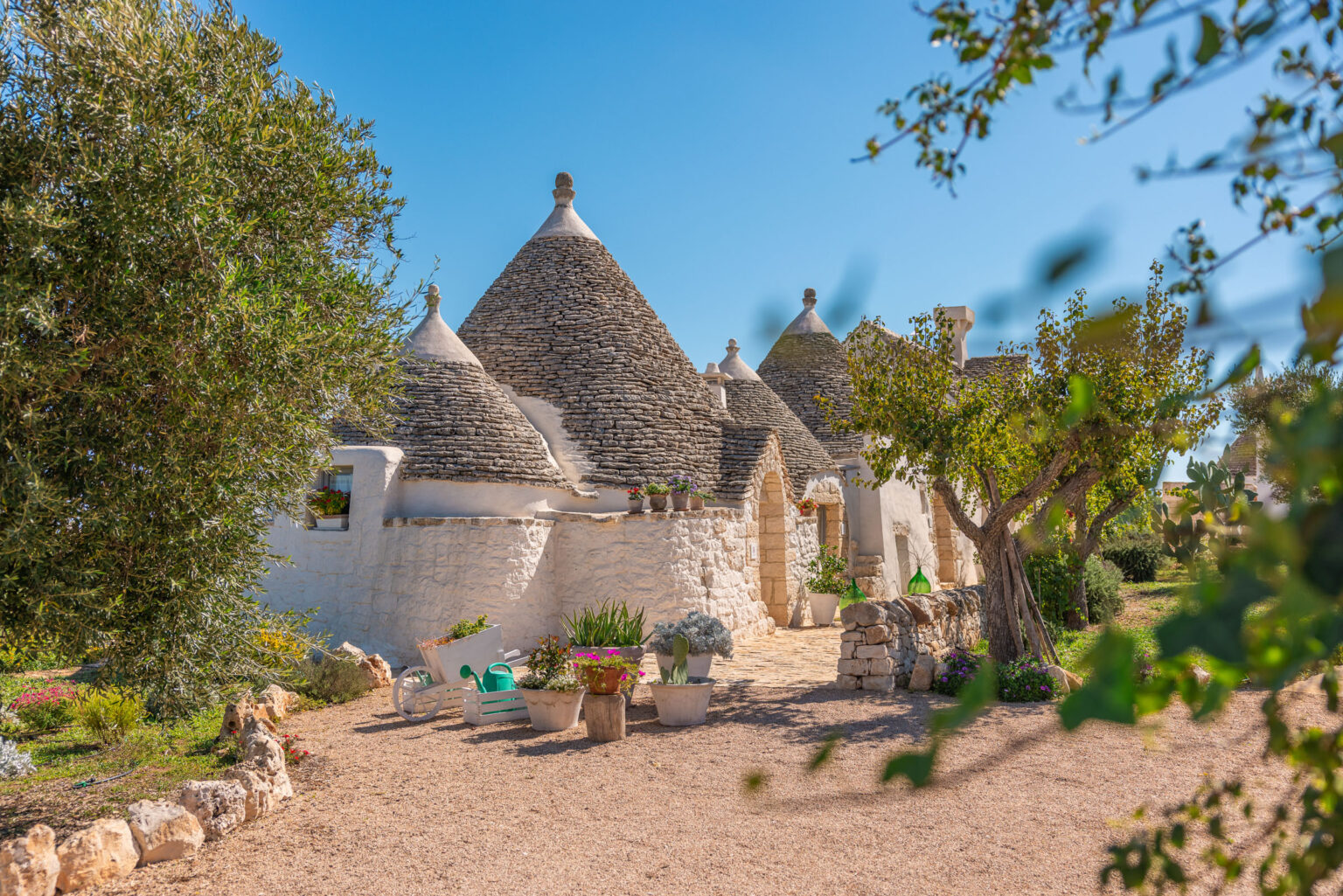 Trulli houses in a scenic landscape, featuring conical stone roofs and whitewashed walls, surrounded by lush greenery and colorful flower pots in a sunny setting. | Only on SopranoVillas