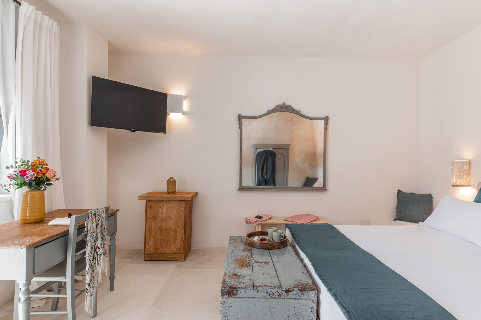 Cozy hotel room featuring a comfortable bed, a rustic wooden table with flowers, a large mirror, and a flat-screen TV, all set in a bright, minimalist design. | Only on SopranoVillas
