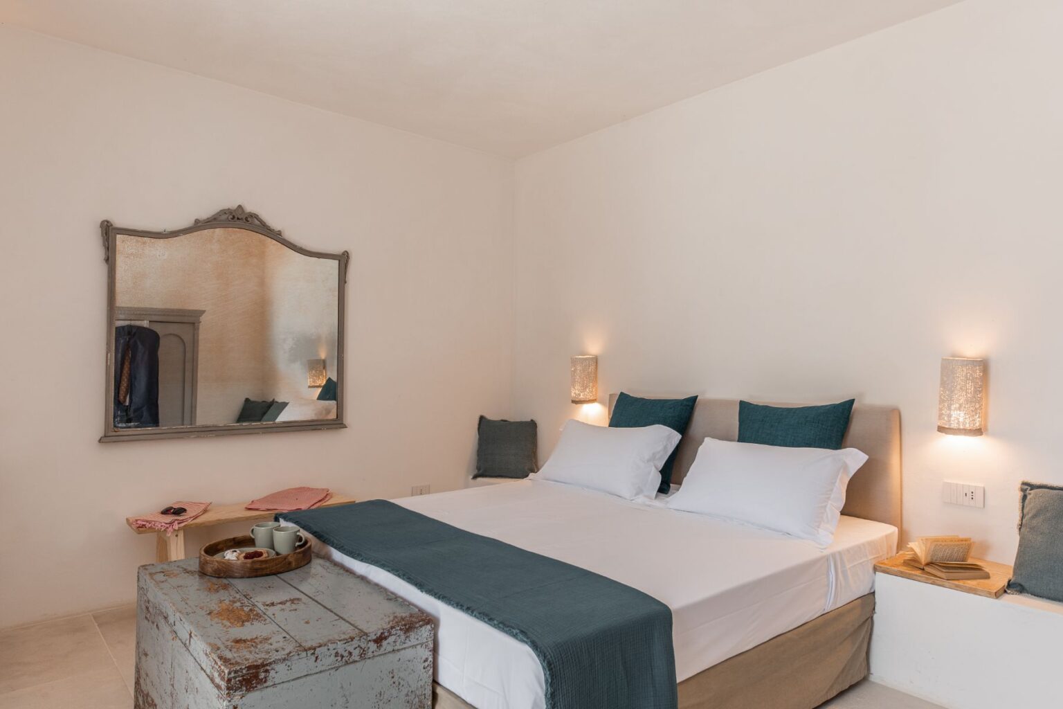 Modern and minimalist bedroom featuring a spacious bed with white linens and teal accents, a vintage-style mirror, and soft lighting from wall sconces, complemented by rustic decor elements. | Only on SopranoVillas