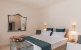 Modern and minimalist bedroom featuring a spacious bed with white linens and teal accents, a vintage-style mirror, and soft lighting from wall sconces, complemented by rustic decor elements.