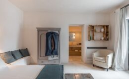 Modern hotel room featuring a minimalist design with a bed, blue decorative pillows, an antique wardrobe with a suit bag, and a cozy armchair. A bathroom is visible in the background, showcasing beige tones and contemporary fixtures. Natural light flows through large windows, enhancing the serene atmosphere.