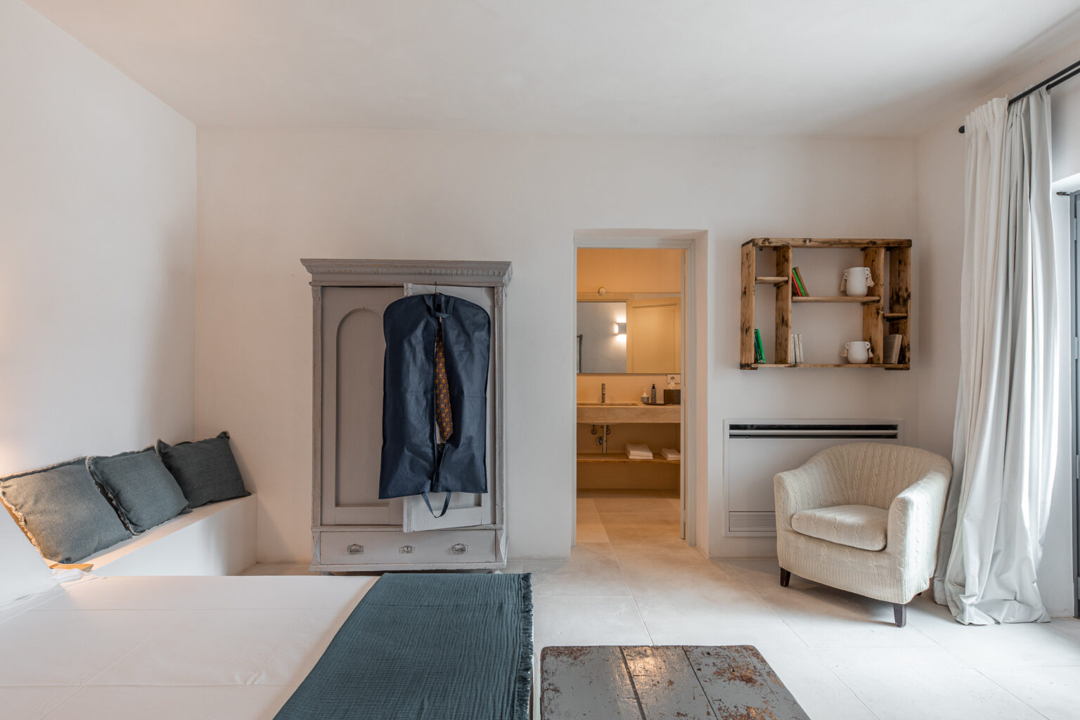 Modern minimalist bedroom featuring a gray wardrobe, a cozy armchair, and an en-suite bathroom entrance. The room has light-colored walls, a neutral bedspread, and stylish decor elements, creating a serene atmosphere. | Only on SopranoVillas