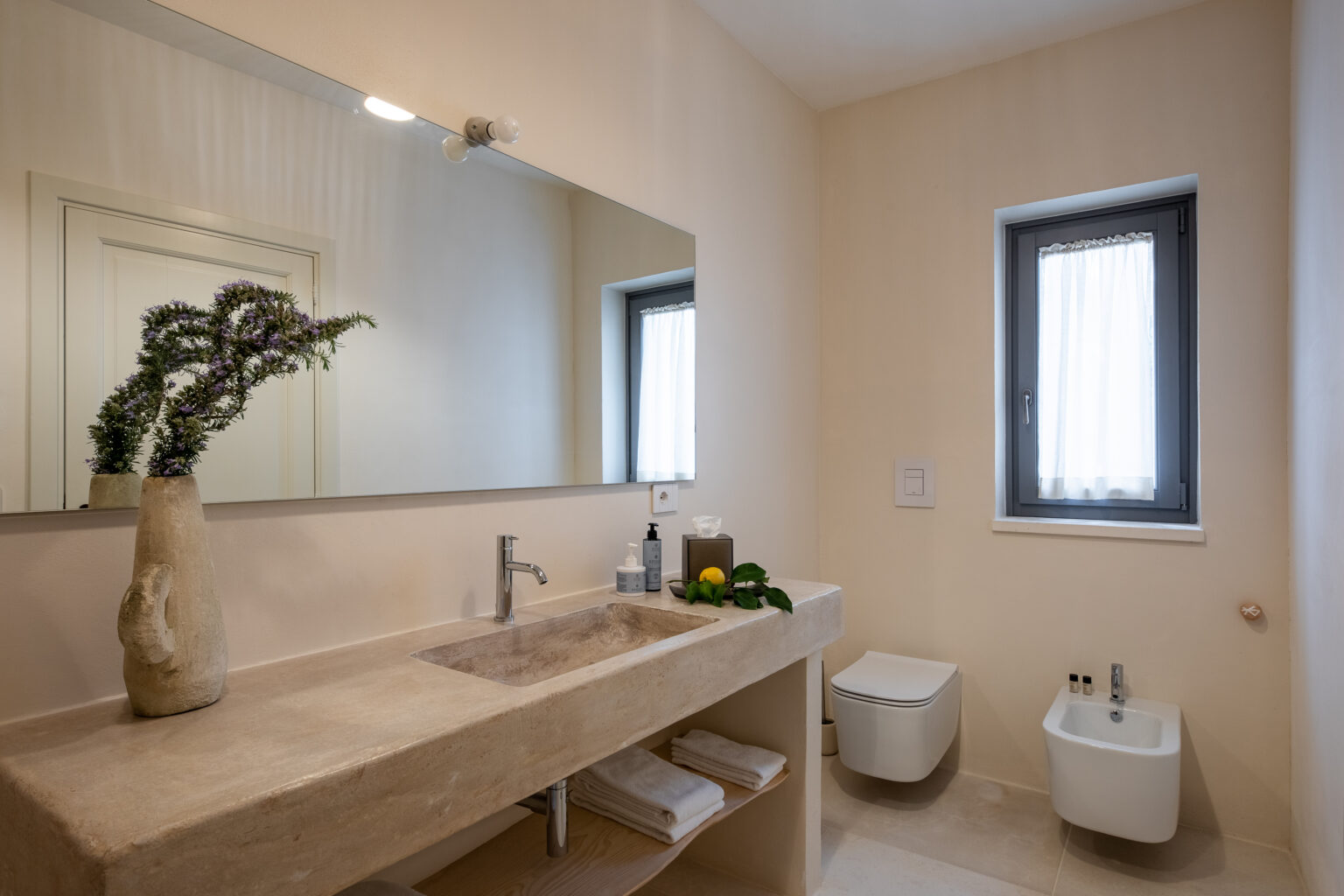 Modern bathroom interior featuring a minimalist design with a natural stone sink, stylish faucet, and a large mirror. Decorated with a vase of fresh greenery and citrus fruits, the space includes a wall-mounted toilet and bidet, enhancing its contemporary aesthetic. Natural light filters through a window with sheer curtains, creating a serene atmosphere. | Only on SopranoVillas