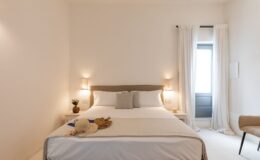 Modern bedroom interior featuring a neatly made bed with neutral linens, decorative pillows, and natural lighting from a nearby window. A small bedside table holds a decorative vase and a candle, while a stylish chair adds to the minimalistic design. Ideal for showcasing contemporary home decor and comfort.