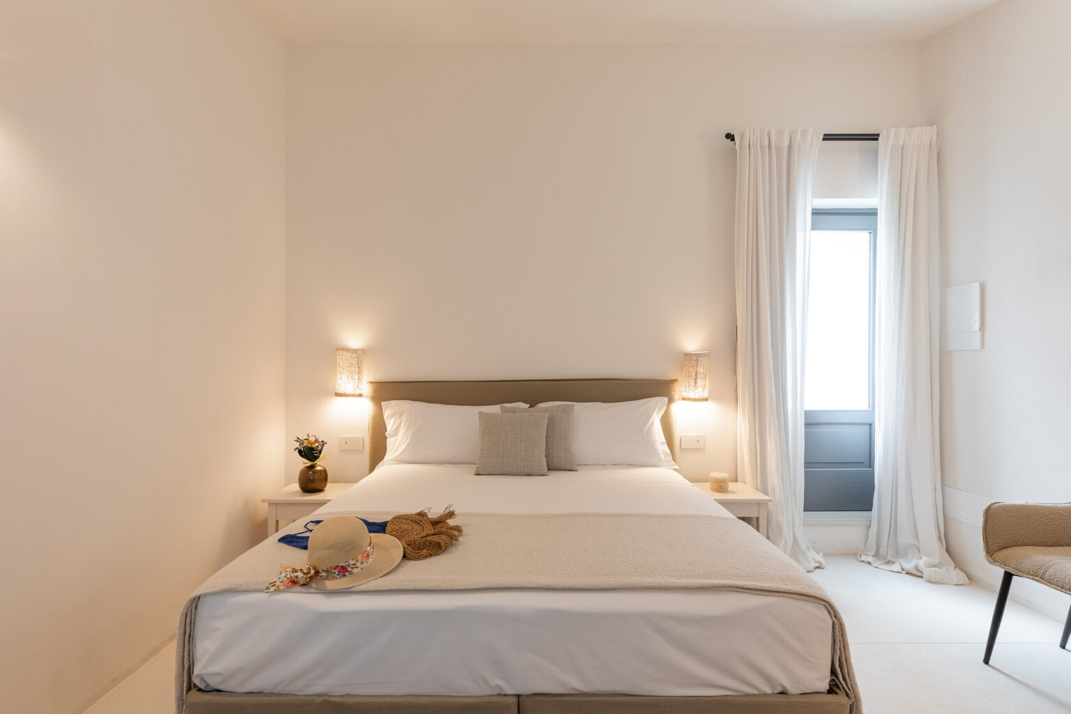 Modern bedroom featuring a comfortable queen-sized bed with soft linens, bedside lamps, and a cozy armchair. Natural light streams through a window with sheer curtains, creating a bright and inviting atmosphere. A plush teddy bear adds a touch of warmth to the decor. | Only on SopranoVillas