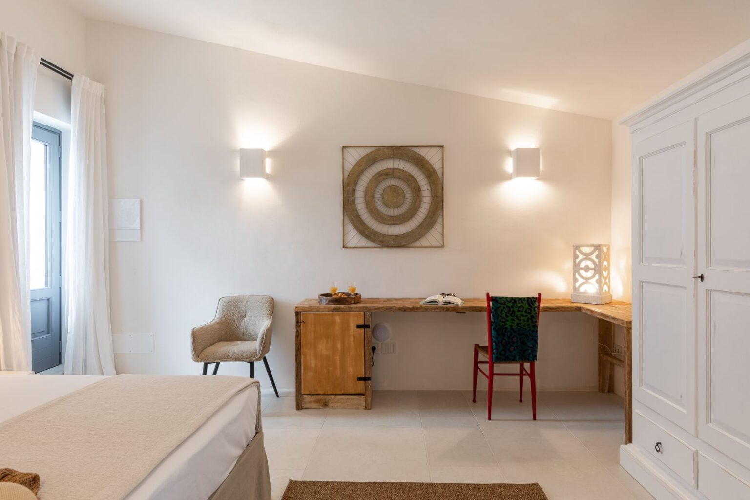 Modern interior design in a cozy bedroom featuring a beige bedspread, a wooden desk with books and drinks, and a colorful chair. Natural light streams in through a window draped in white curtains, complemented by stylish wall sconces and decorative wall art. | Only on SopranoVillas