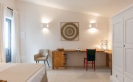 Modern interior design in a cozy bedroom featuring a beige bedspread, a wooden desk with books and drinks, and a colorful chair. Natural light streams in through a window draped in white curtains, complemented by stylish wall sconces and decorative wall art.