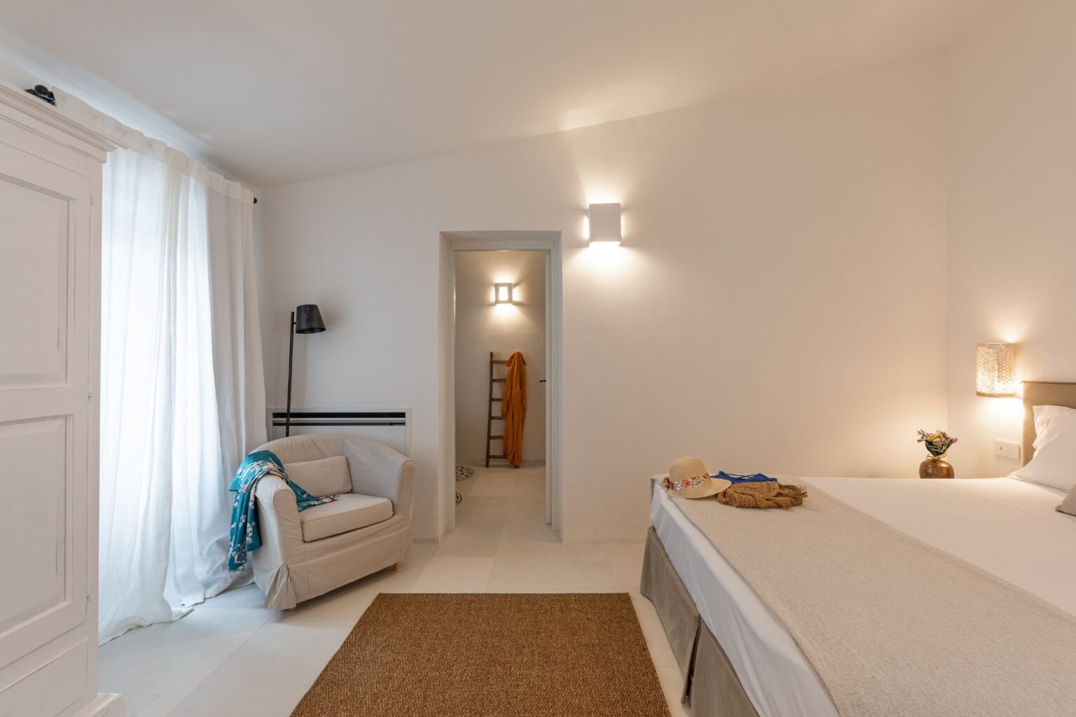 Cozy bedroom featuring a comfortable bed with a neutral color scheme, light-colored walls, and natural lighting. A soft rug on the floor complements the space, while a plush armchair and decorative throw add warmth. A doorway leads to a modern bathroom, enhancing the serene atmosphere. | Only on SopranoVillas