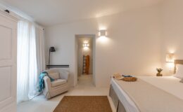 Cozy bedroom featuring a comfortable bed with a neutral color scheme, light-colored walls, and natural lighting. A soft rug on the floor complements the space, while a plush armchair and decorative throw add warmth. A doorway leads to a modern bathroom, enhancing the serene atmosphere.