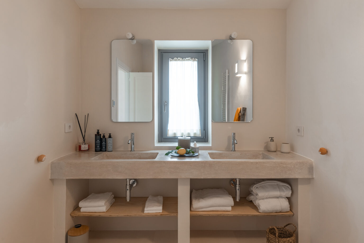 Modern bathroom design featuring a spacious concrete vanity with dual sinks, two mirrors, and ample storage space underneath. Natural light filters through a window, enhancing the minimalist aesthetic. Neatly arranged towels and decorative items complete the serene atmosphere. | Only on SopranoVillas