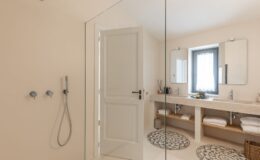 Modern bathroom featuring a spacious glass shower, dual sinks, and elegant decor. The light-colored walls and minimalist design create a serene atmosphere. Natural light streams in through a window with sheer curtains, enhancing the overall ambiance. Soft towels are neatly arranged on a wooden shelf, complemented by stylish bath accessories.